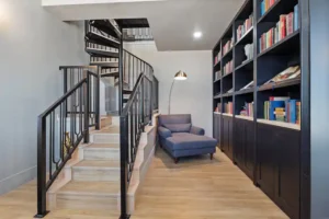 View of a custom build library with black shelves, a sofa chair, and a spiral staircase.
