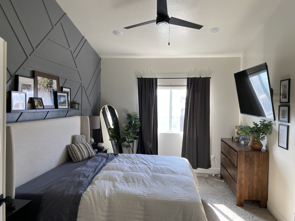 "A modern bedroom with a dark geometric accent wall behind a large upholstered bed. The room features neutral tones, a wooden dresser with a mounted TV above it, dark curtains covering a window, and a ceiling fan. A round mirror with a gold frame leans against the wall next to a green potted plant. The bed is adorned with a white and dark gray comforter and decorative pillows.