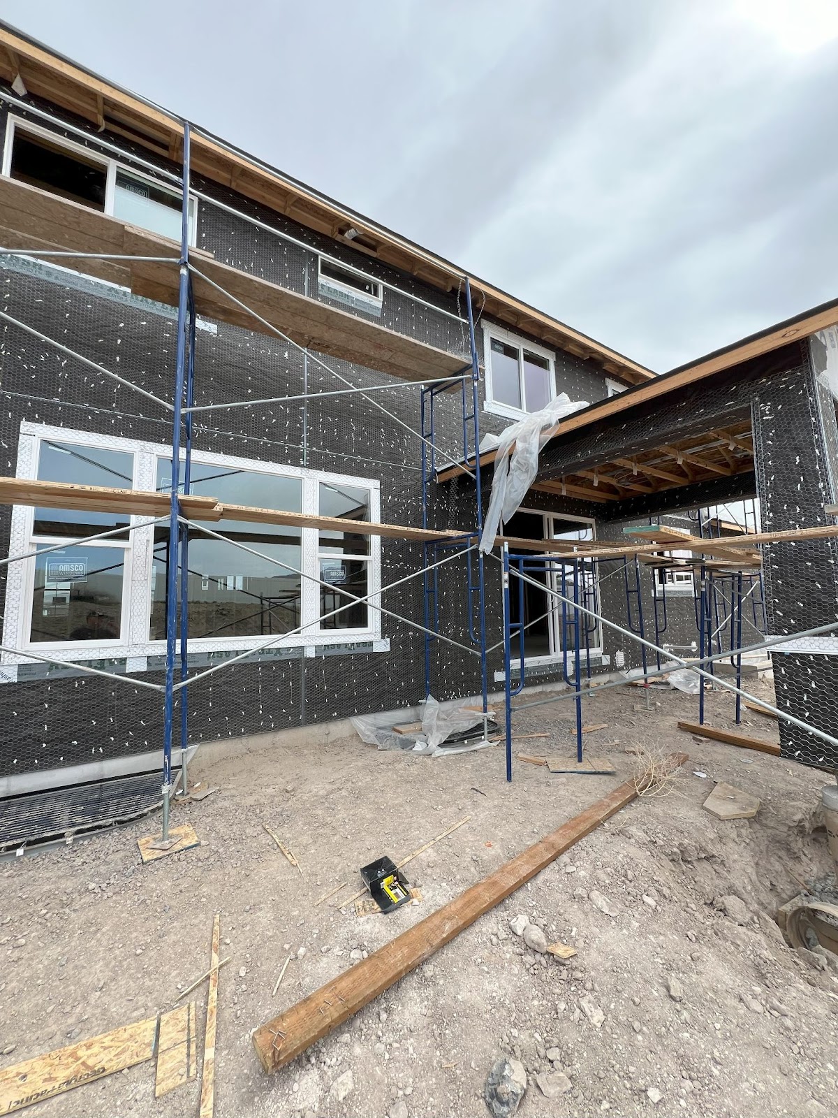 Under-construction house with black weather barrier, newly installed windows, and scaffolding.