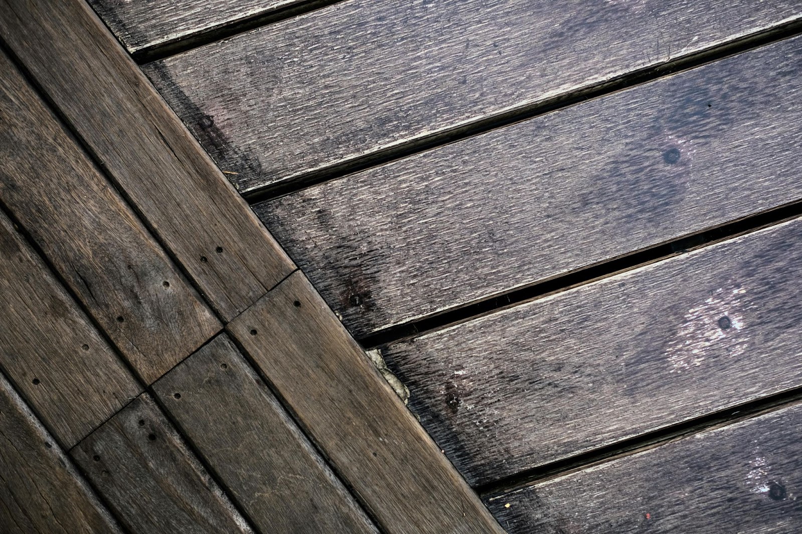 Two sections of weathered wooden deck planks meet at a diagonal angle, showing individual boards and grain.