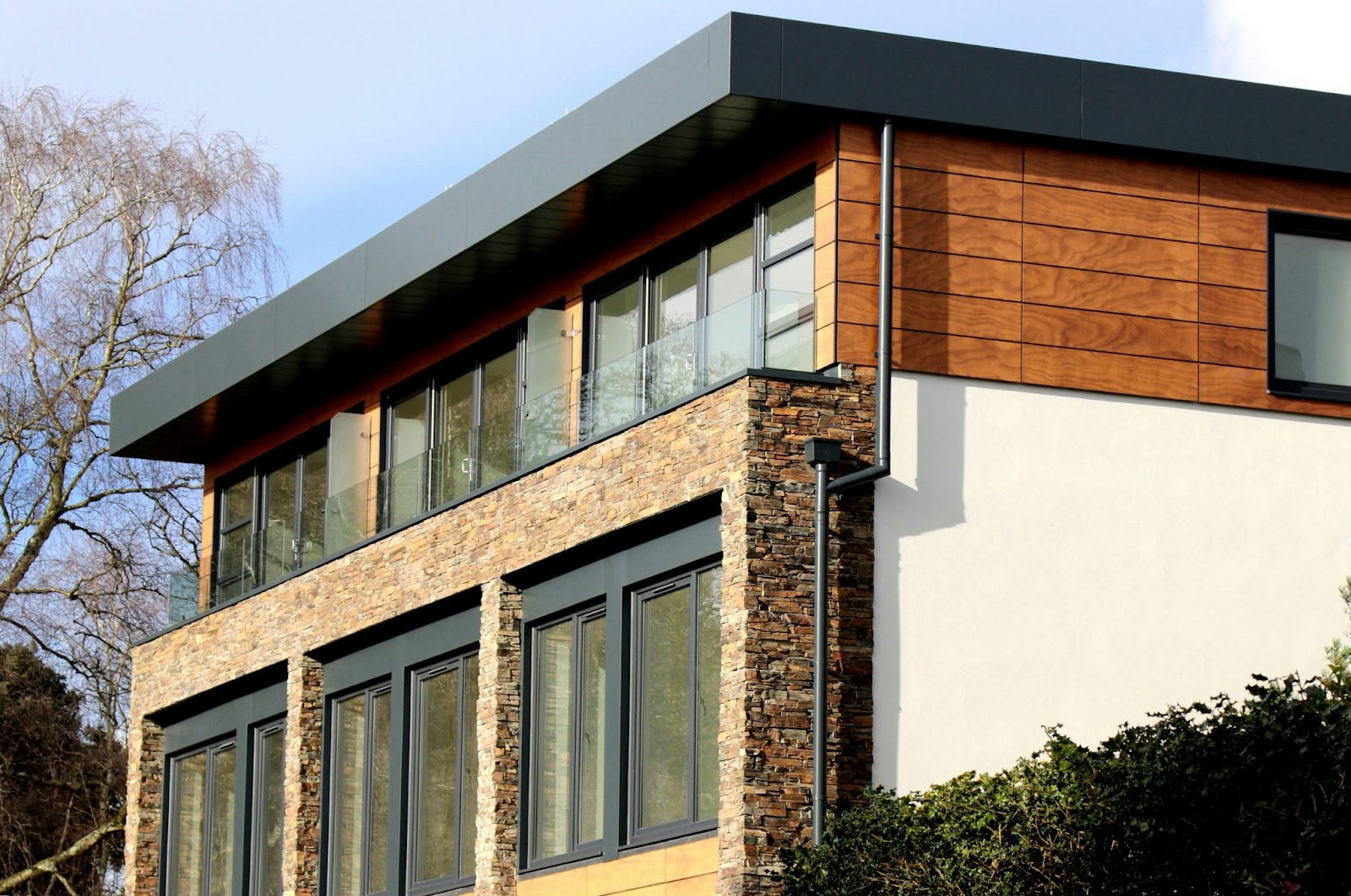 A modern home with a dark roofline features a mix of wood paneling, stone facade, and white stucco walls with large windows.