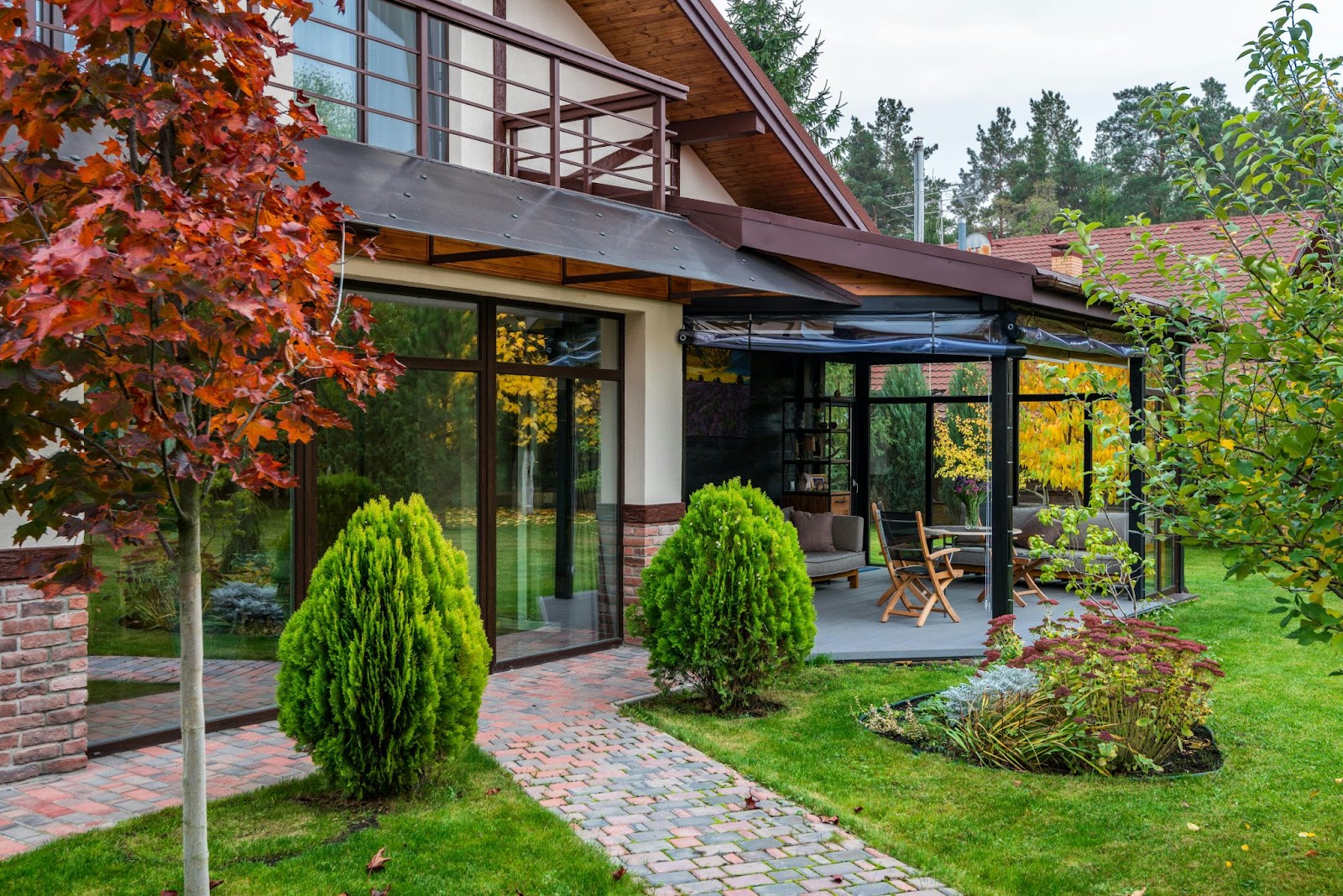 A modern house features large glass windows and an outdoor patio surrounded by vibrant green grass and autumn-colored trees.