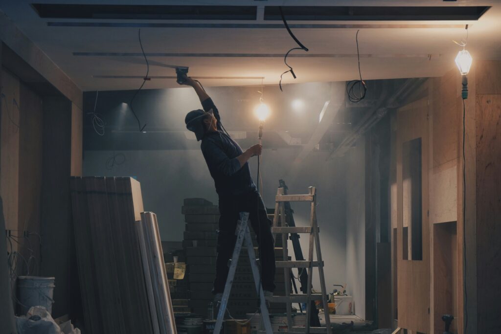 Home renovation construction work - electrician installing wiring on ladder in unfinished room with exposed beams and construction dust
