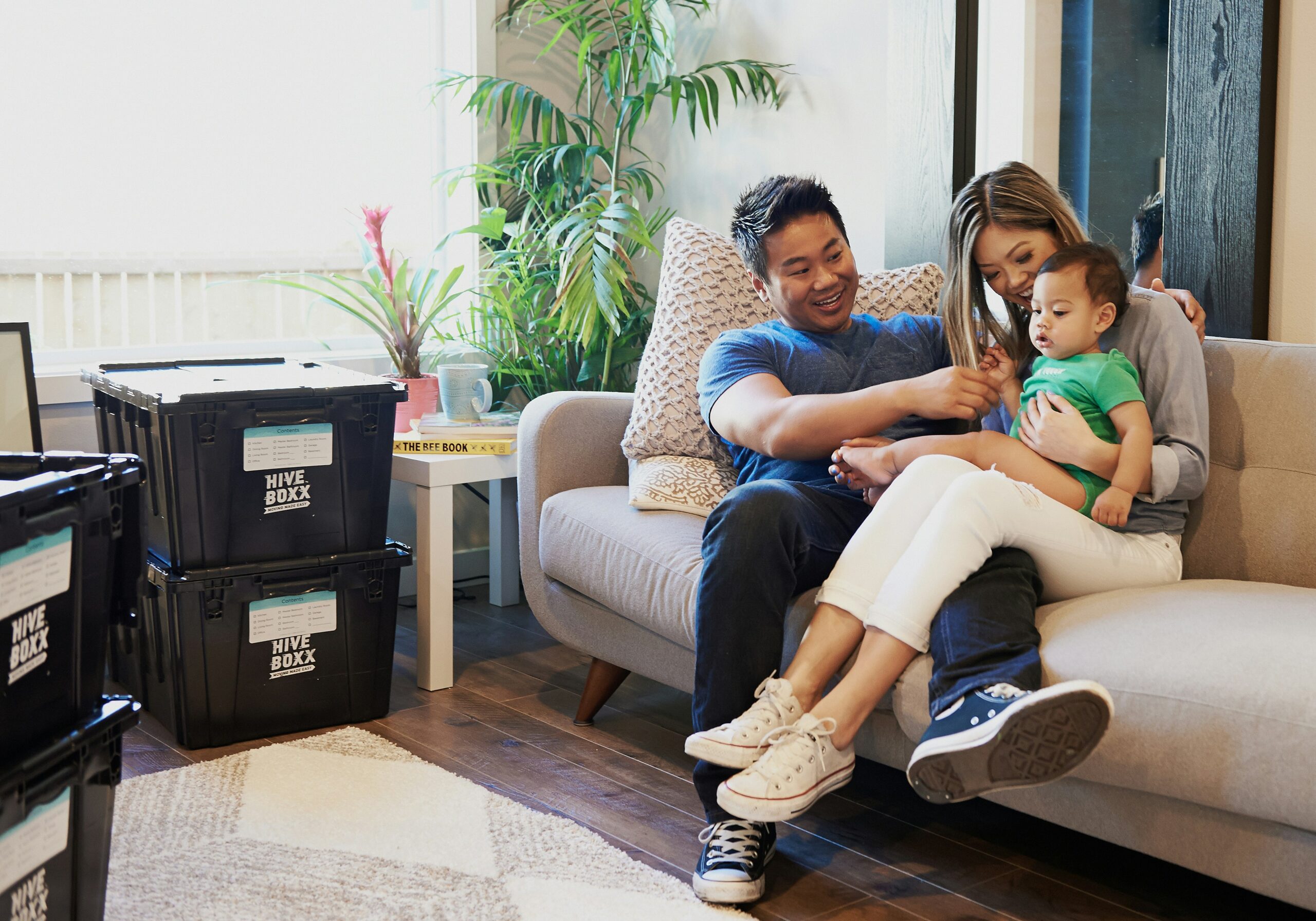 Smiling parents with young child sitting on a couch surrounded by moving boxes in their freshly built home
