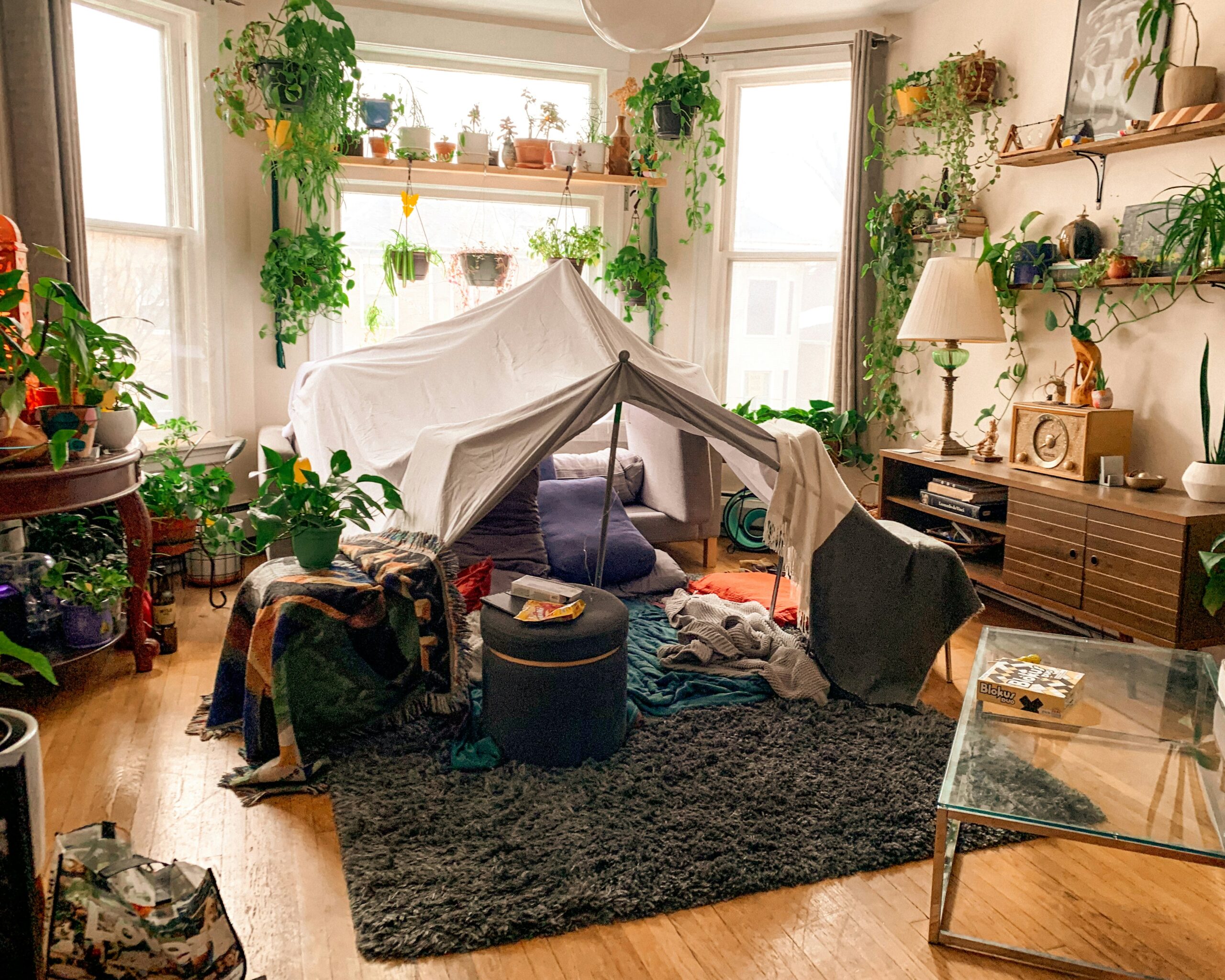 Indoor living room filled with natural light and lush green plants, featuring a cozy blanket fort in the center made from white sheets and surrounded by pillows and blankets, creating a playful and inviting space.