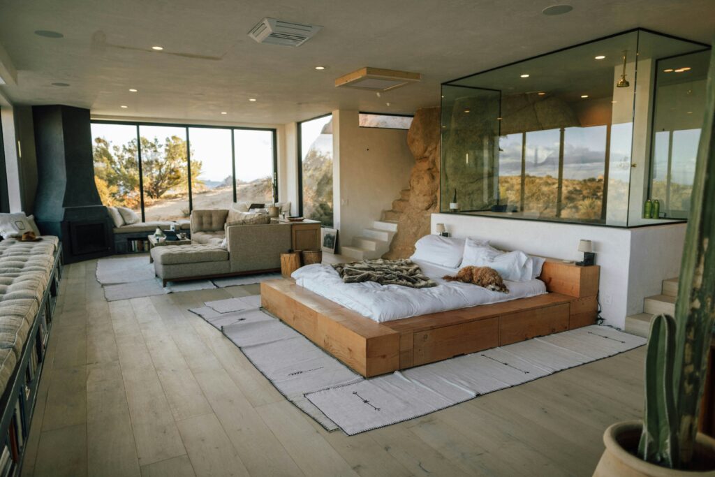 Modern desert home interior featuring an open-plan bedroom and living area with floor-to-ceiling windows, natural wood platform bed, glass-enclosed bathroom, and panoramic mountain views