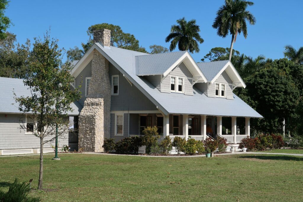 Traditional farmhouse style home exterior with metal roof, stone chimney, front porch, and tropical landscaping in residential neighborhood