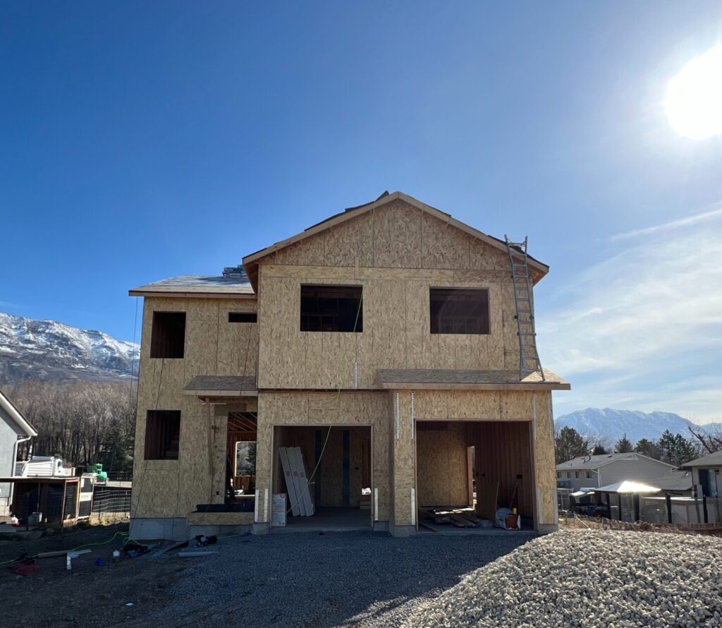 A two-story custom home in Utah under construction, framed with plywood and surrounded by gravel, captured on a clear blue day with mountains in the background.