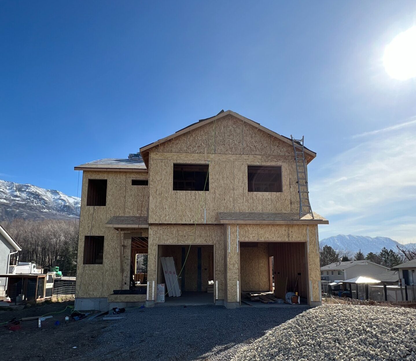 A two-story custom home in Utah under construction, framed with plywood and surrounded by gravel, captured on a clear blue day with mountains in the background.