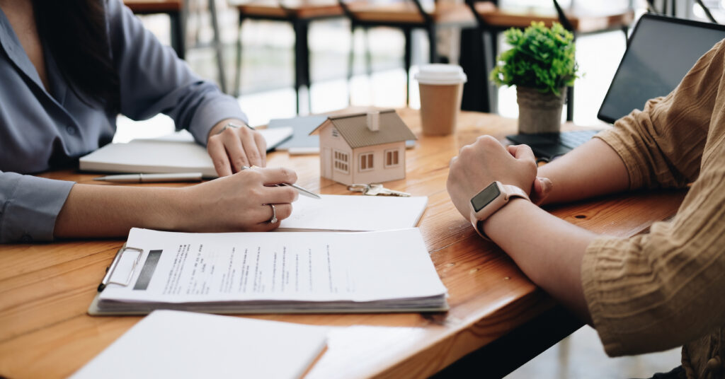 Two people meeting at a table to discuss real estate contracts with a small house model and paperwork, representing investment property planning.
