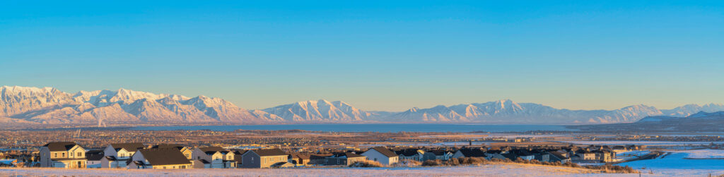 A beautiful panoramic view of a new home community in Saratoga Springs, Utah, with snow-covered mountains and Utah Lake in the background under a clear blue sky.