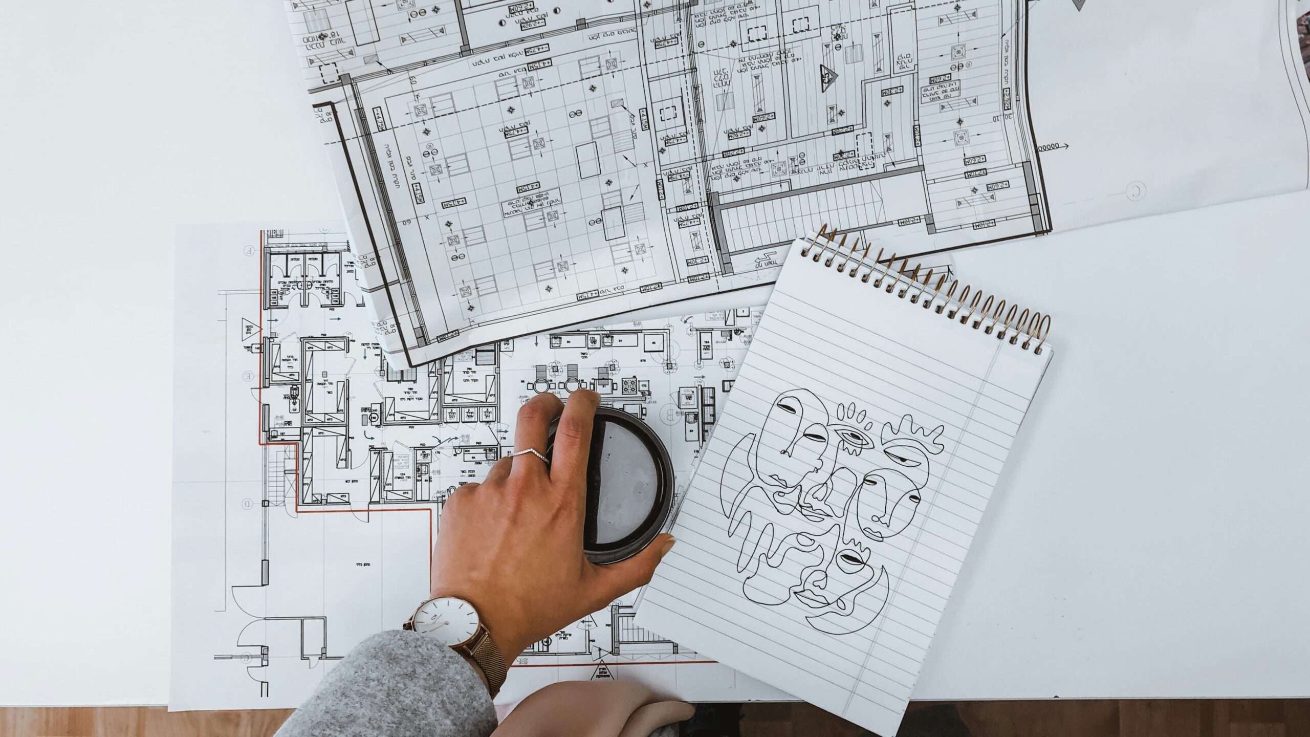 A person reviewing detailed architectural floor plans for a custom home with a notebook and coffee cup on the desk, symbolizing early design planning.