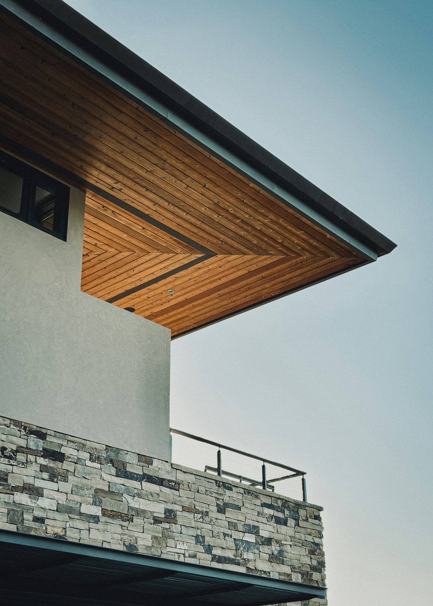 Modern custom home exterior detail featuring warm wood soffit, dark metal roofline, and stacked stone accents — a 2025 Utah exterior design trend
