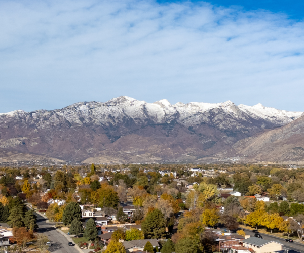 A panoramic view of American Fork, Utah, featuring a suburban neighborhood with colorful autumn foliage in the foreground and the snow-capped Wasatch Mountains in the background under a blue sky with scattered clouds.